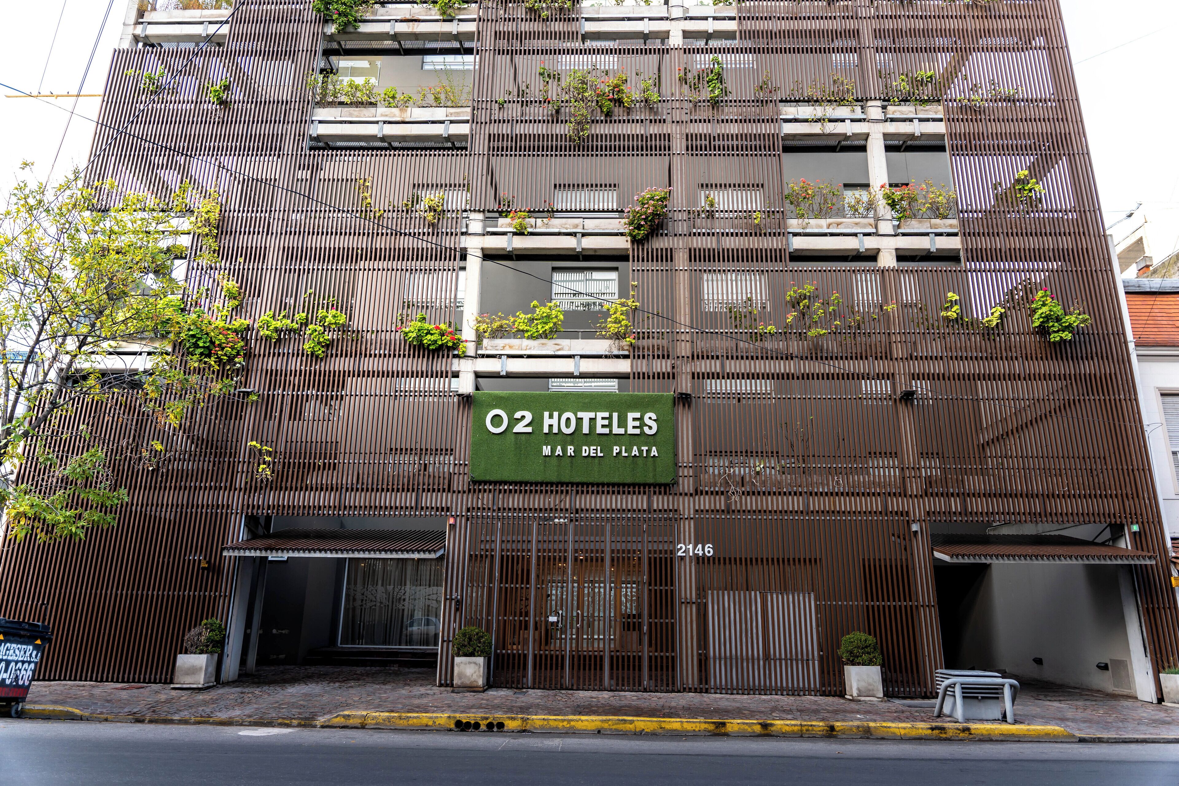 Fachada de hotel con diseño de panel metálico vertical, balcones con plantas y un letrero que dice "O2 HOTELES MAR DEL PLATA 2146".