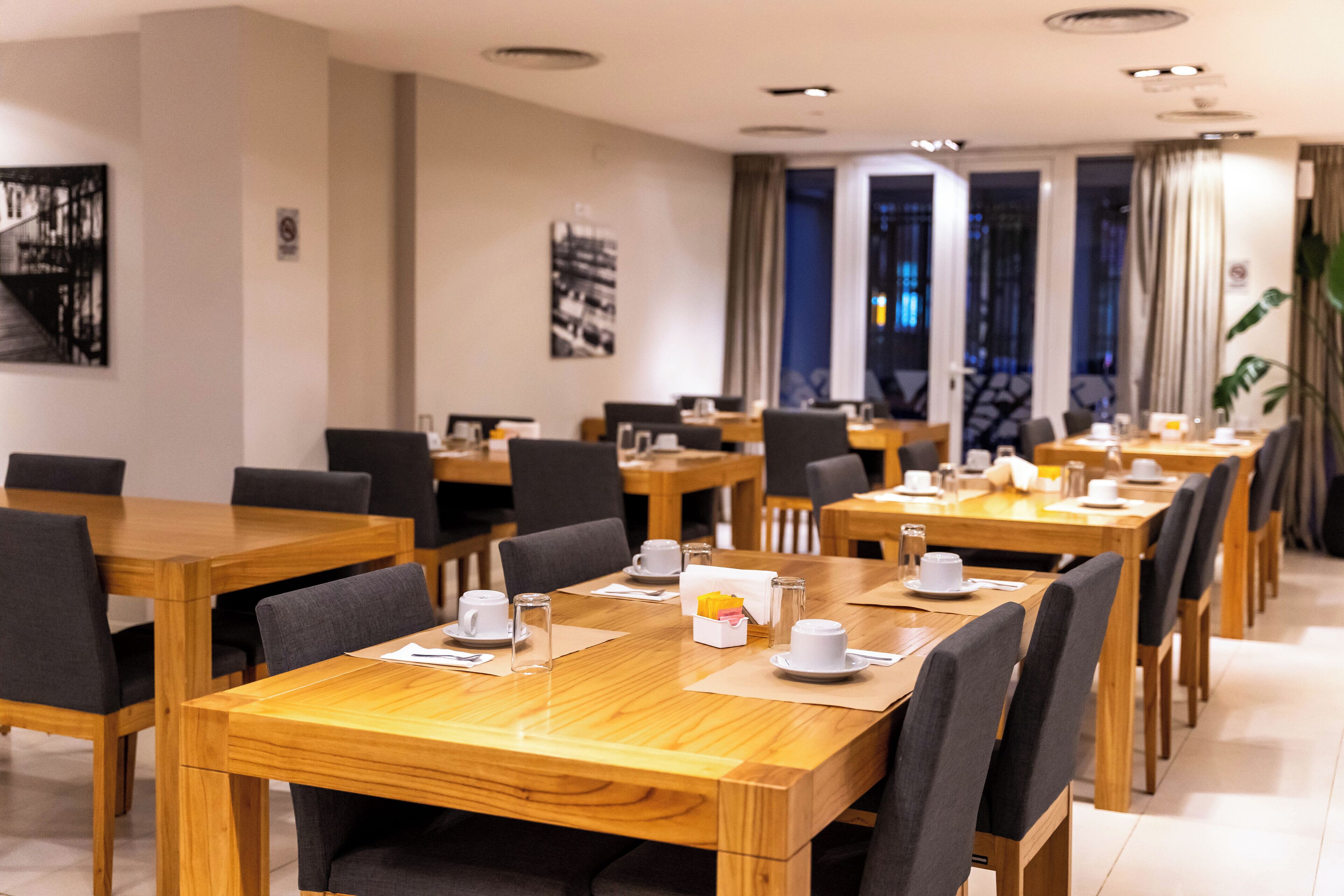 A modern dining area with wooden tables and black chairs, equipped with white cups and saucers.