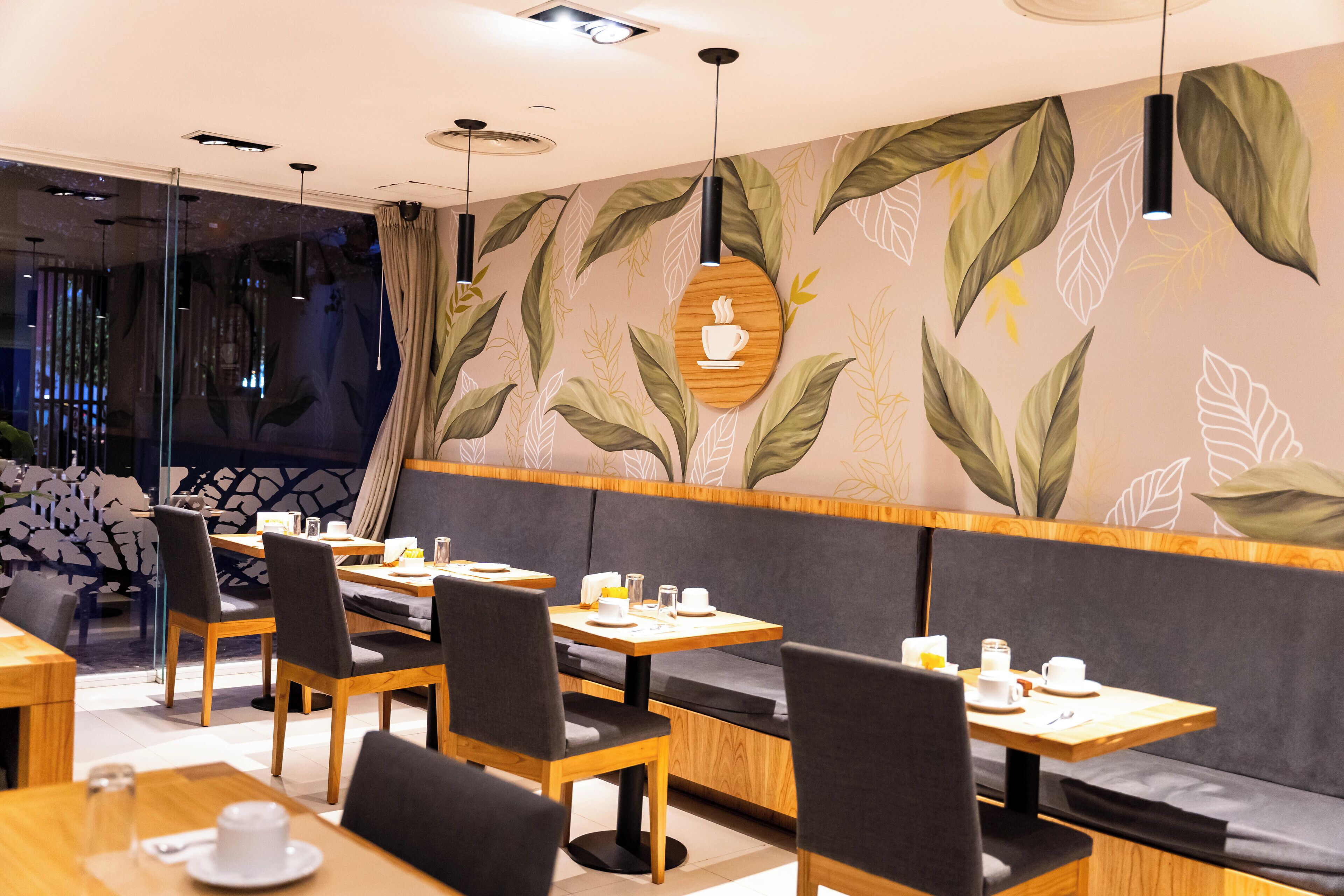 A modern cafe with wooden tables and black chairs, featuring a leafy wall mural and pendant lighting.