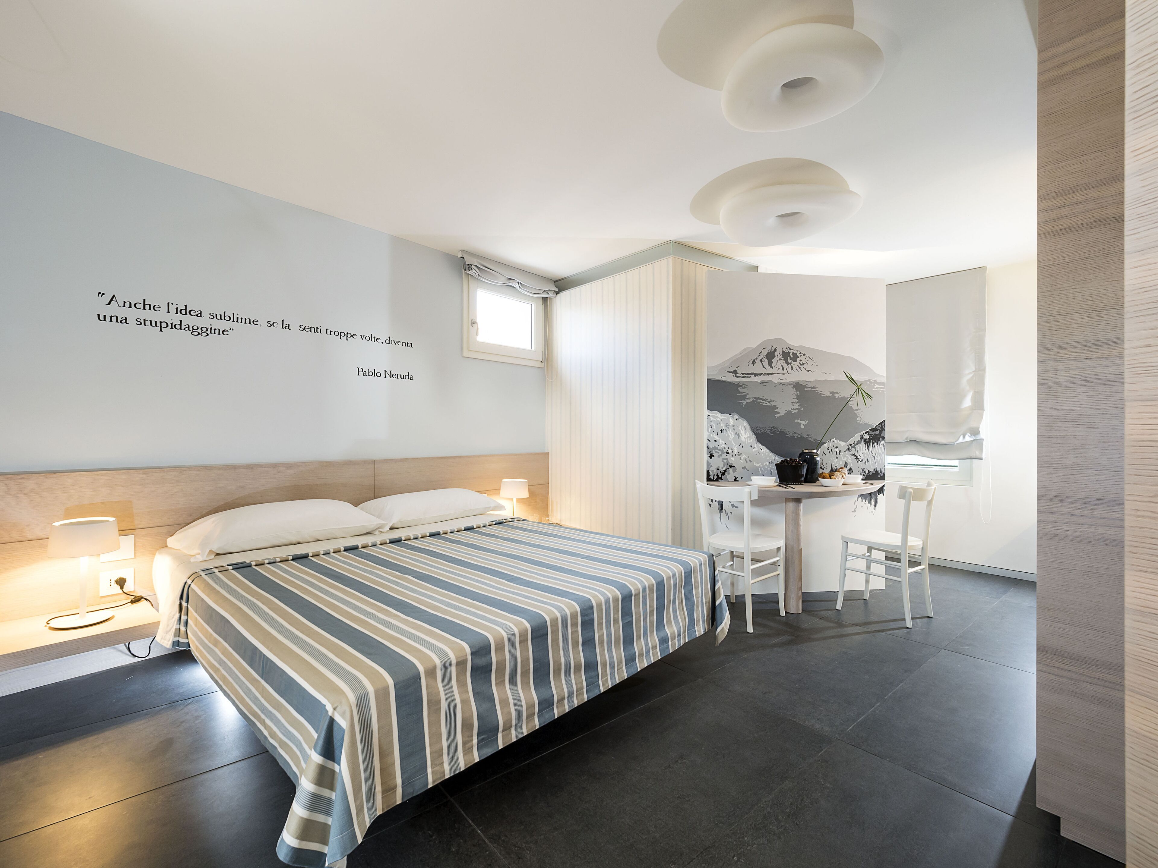 Een moderne slaapkamer met een bed, nachtlampjes, een bureau met stoelen en een muur met een citaat.