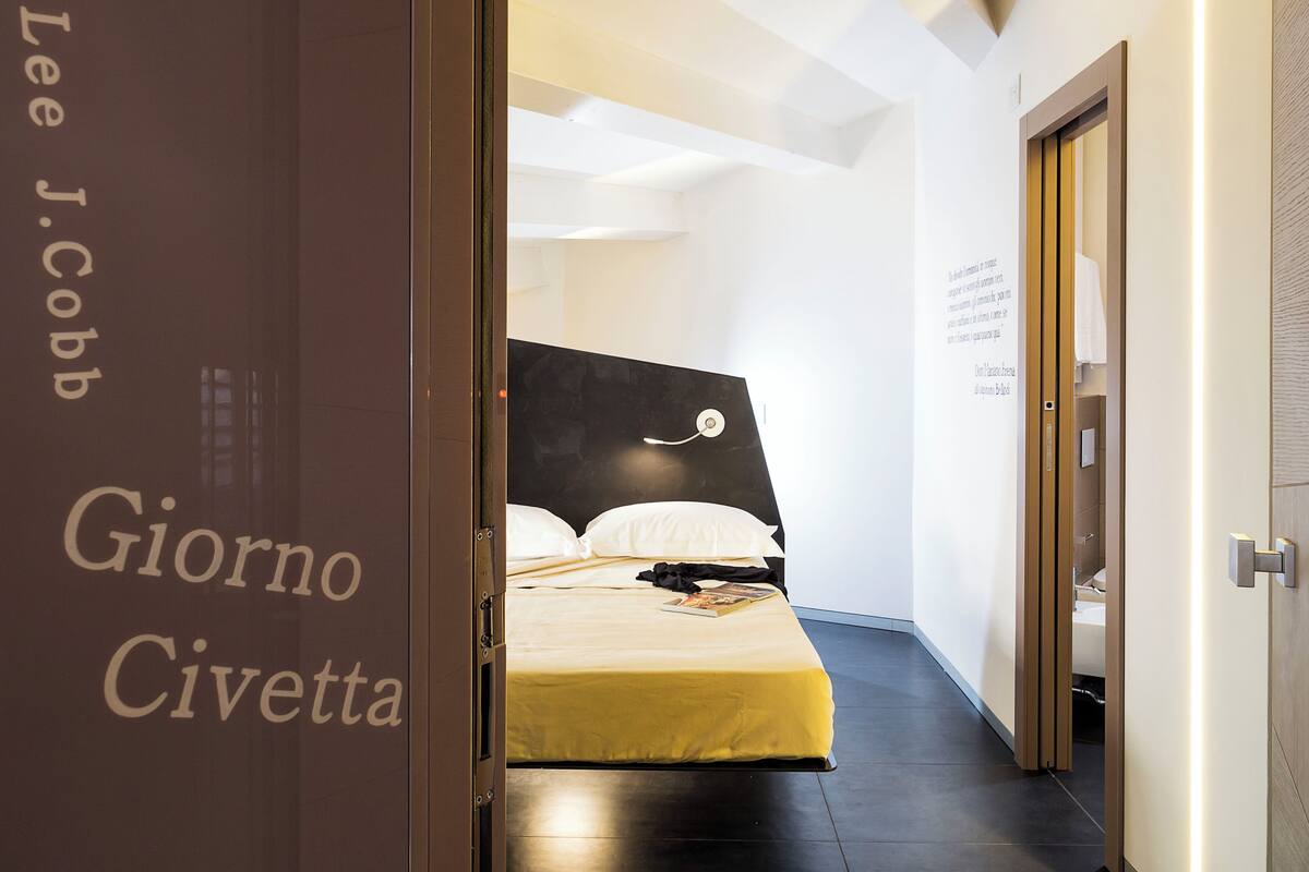 A hotel room with a bed, a bedside table, and a nightstand. There is a door with a sign that reads "Lee J. Cobb Giorno Civetta". The room has a modern design with a white ceiling and walls.