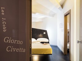 Una camera d'albergo con un letto, un comodino e una abat-jour. Si vede una porta con un cartello che riporta la scritta 'Lee J. Cobb Giorno Civetta'. La stanza presenta un design moderno, con soffitto e pareti bianchi.