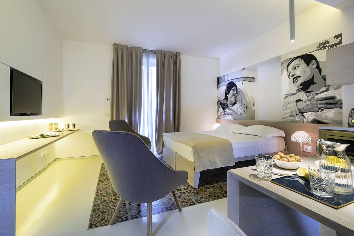 A modern hotel room with a bed, a desk, a chair, a TV, and a dining area with a table and chairs.