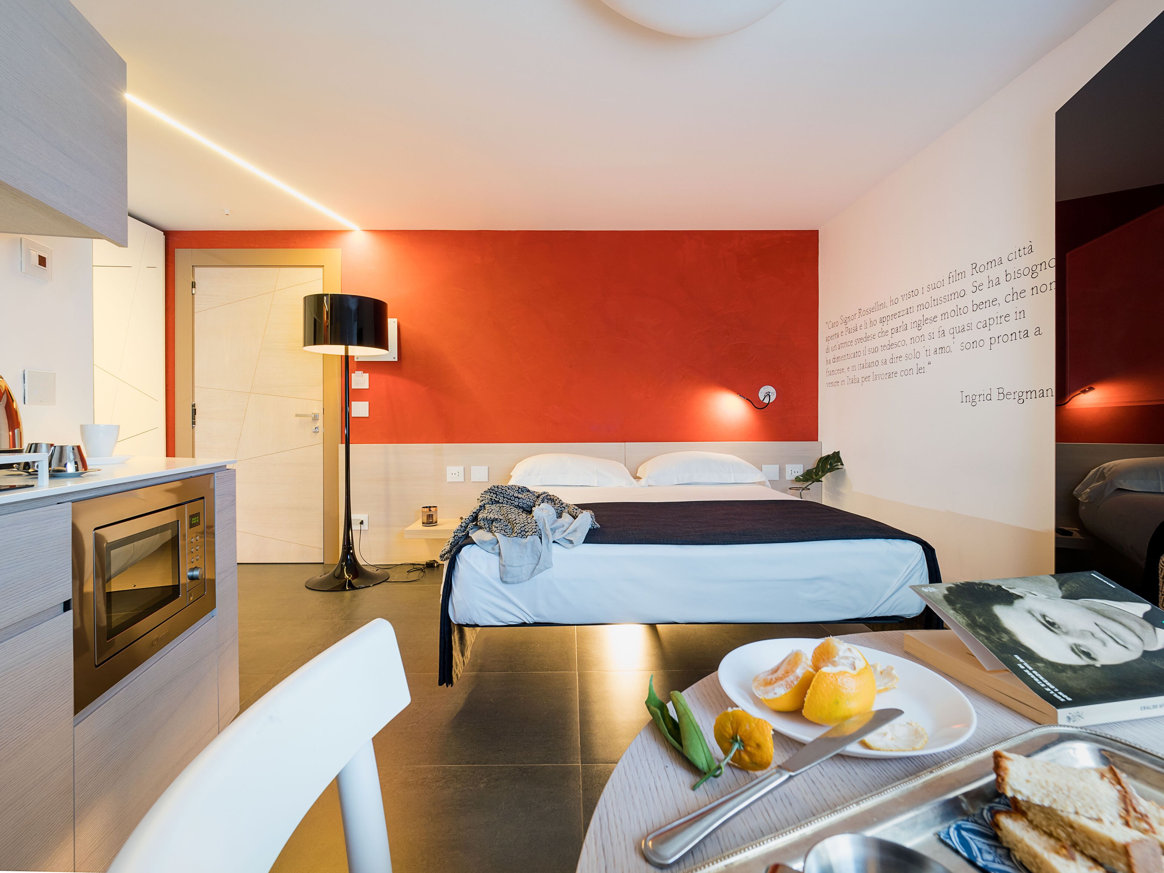 A modern hotel room with a bed, bedside table, and a dining area with a plate of food and a book.