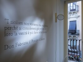 Una pared con una cita en italiano, una ventana que ofrece vista a un edificio y una puerta con pomo circular.