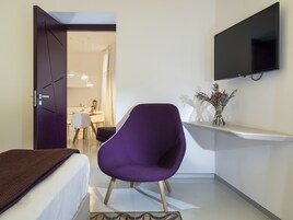 Una habitación pequeña y moderna con una cama, una silla morada, un televisor montado en la pared y una mesita con un florero.
