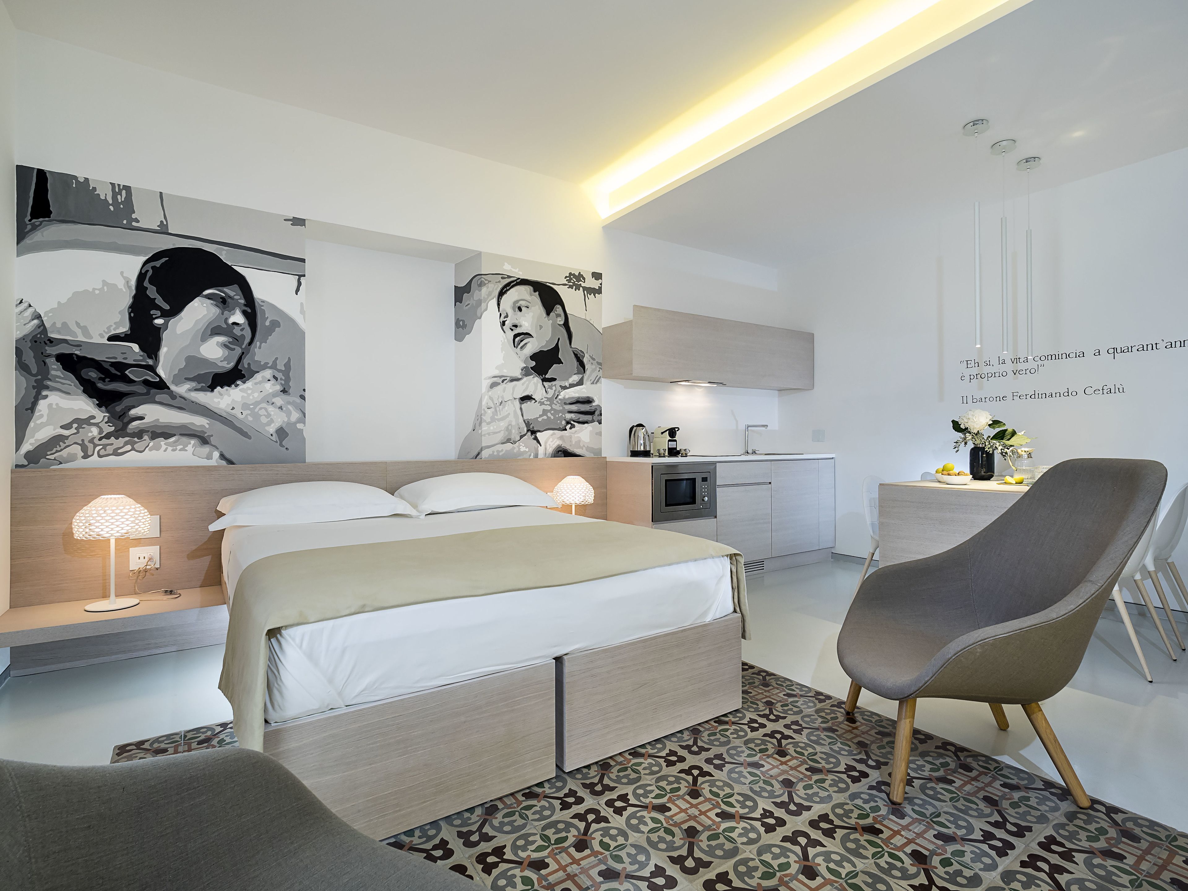 Ein modernes Hotelzimmer mit Bett, Nachttisch, Lampe und einem Sessel. Es gibt eine Küchenzeile mit Mikrowelle und Kaffeemaschine.