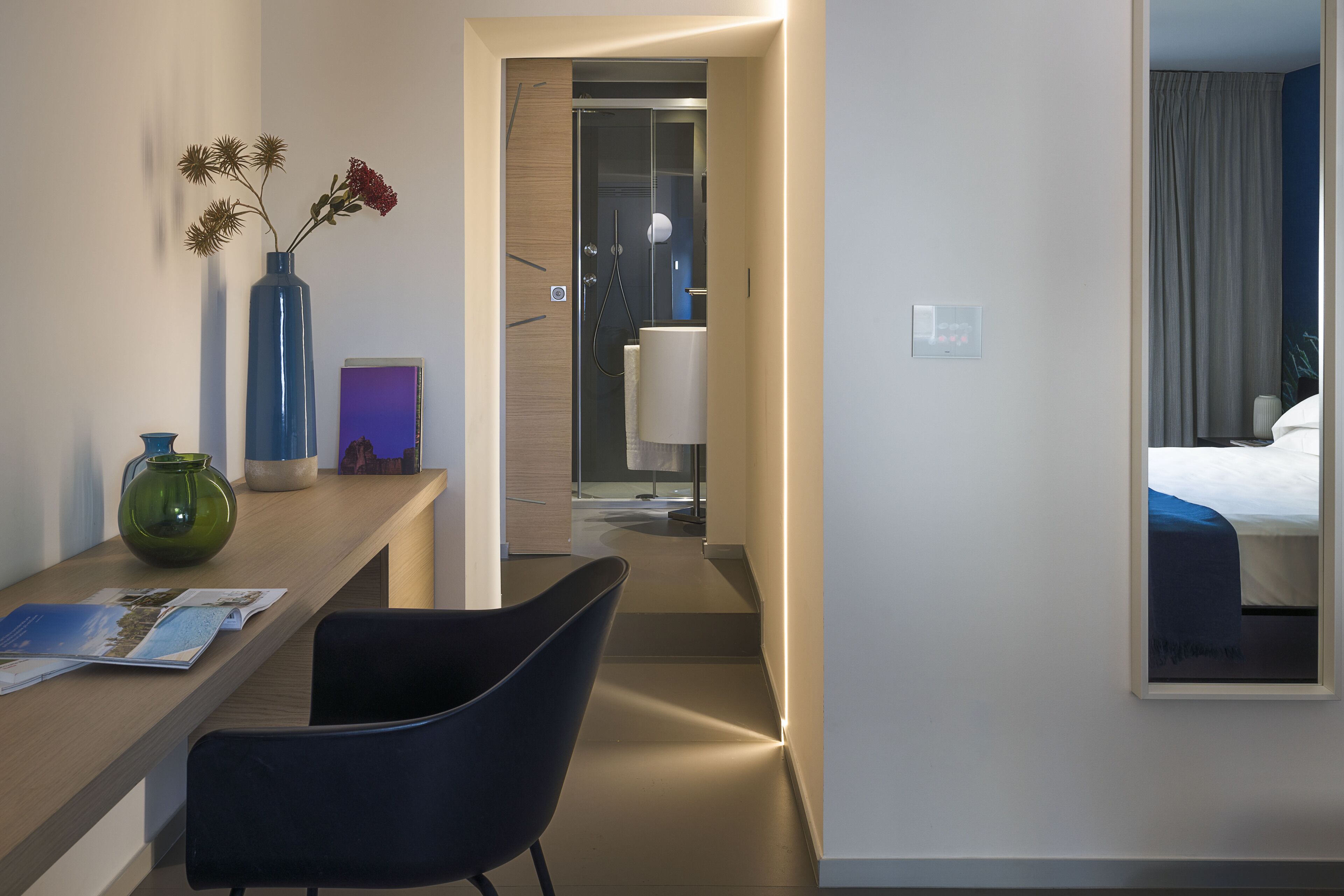 Una camera d'albergo con una scrivania in legno, una sedia, un vaso con fiori secchi, un quadro incorniciato e un letto con copriletto blu.