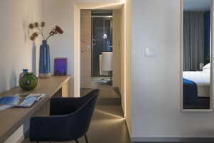 Una camera d'albergo con una scrivania in legno, una sedia, un vaso con fiori secchi, un quadro incorniciato e un letto con copriletto blu.