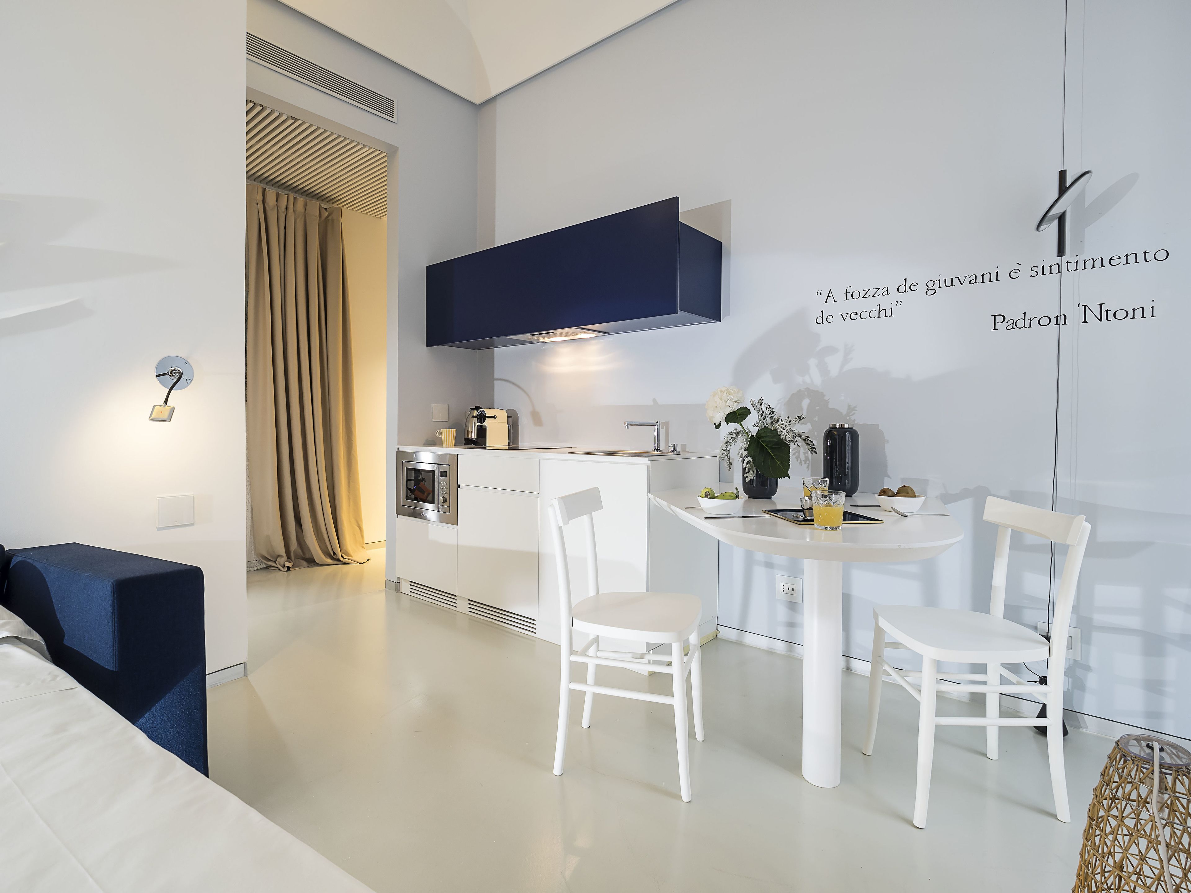 Une cuisine moderne avec des meubles blancs, un four encastré et une hotte de couleur bleu foncé. On y trouve une table à manger avec des chaises blanches et une petite table d’appoint sur laquelle sont posés un verre et une plante.