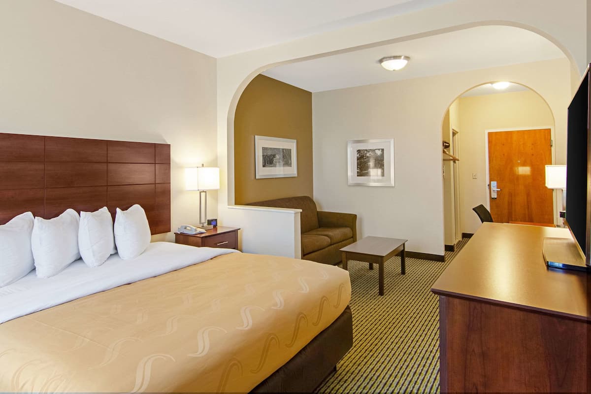 A hotel room with a large bed, a desk, a chair, and a small table. There are two framed pictures on the wall and a wooden door.