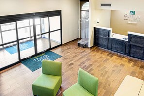 A hotel reception area with a reception desk, two green armchairs, a blue mat with text, and a large window with a view of the outside.