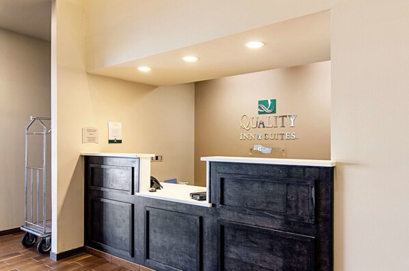 Un mostrador de recepción con detalles de madera oscura y una encimera blanca. Se observa un logotipo sobre el mostrador que dice “QUALITY INN & SUITES”. Un carro metálico está ubicado a la izquierda del mostrador.