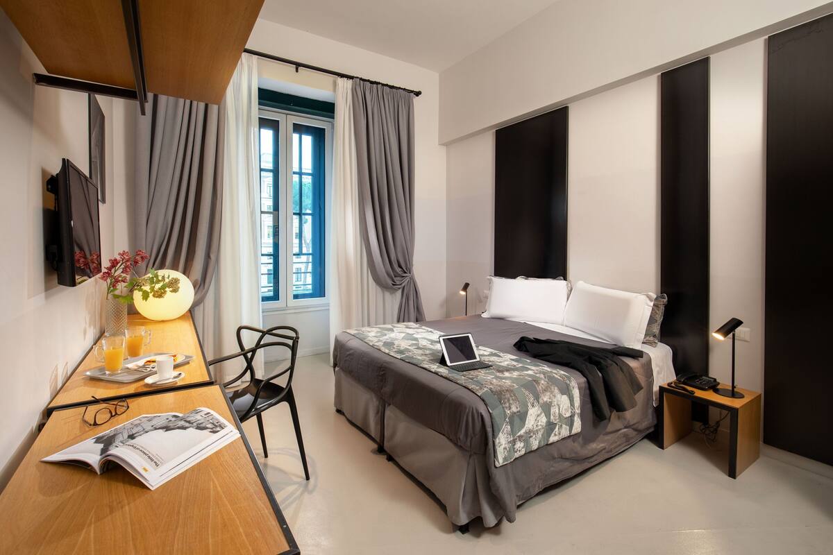 Une chambre moderne avec un lit, un bureau avec un ordinateur portable, une chaise, une table de chevet avec une lampe et une fenêtre avec des rideaux.