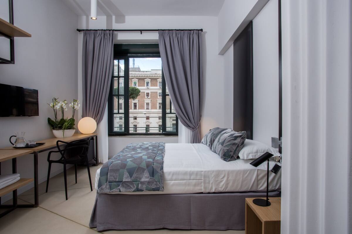 Une chambre moderne avec un lit, un bureau et une chaise, une fenêtre avec des rideaux et une vue sur les bâtiments à l’extérieur.