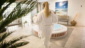 A person in a white robe walking towards a hot tub in a well-lit indoor space with a beach-themed painting on the wall.