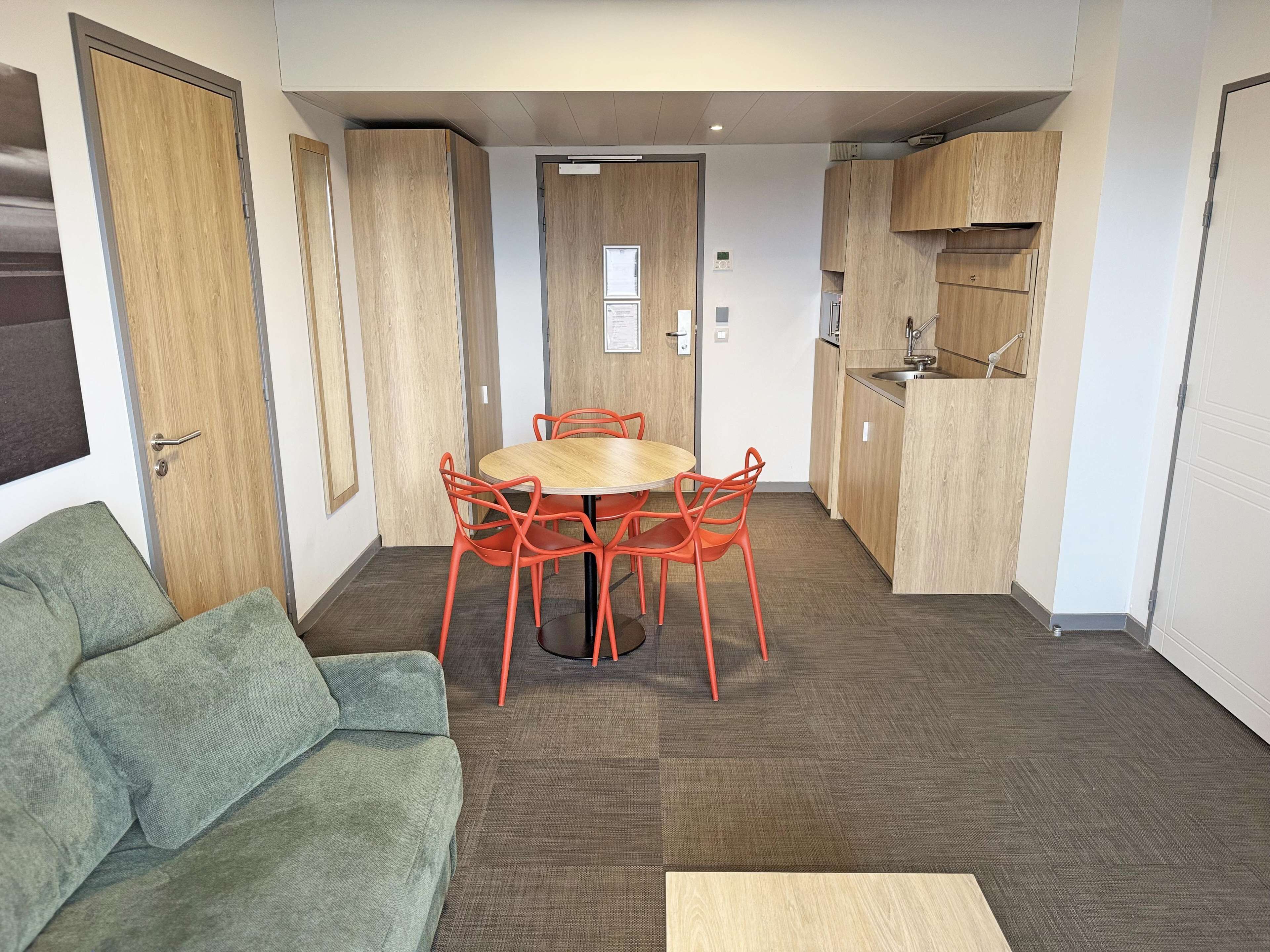A compact living space with a round wooden table and orange chairs, a green sofa, and a kitchenette with wooden cabinets.