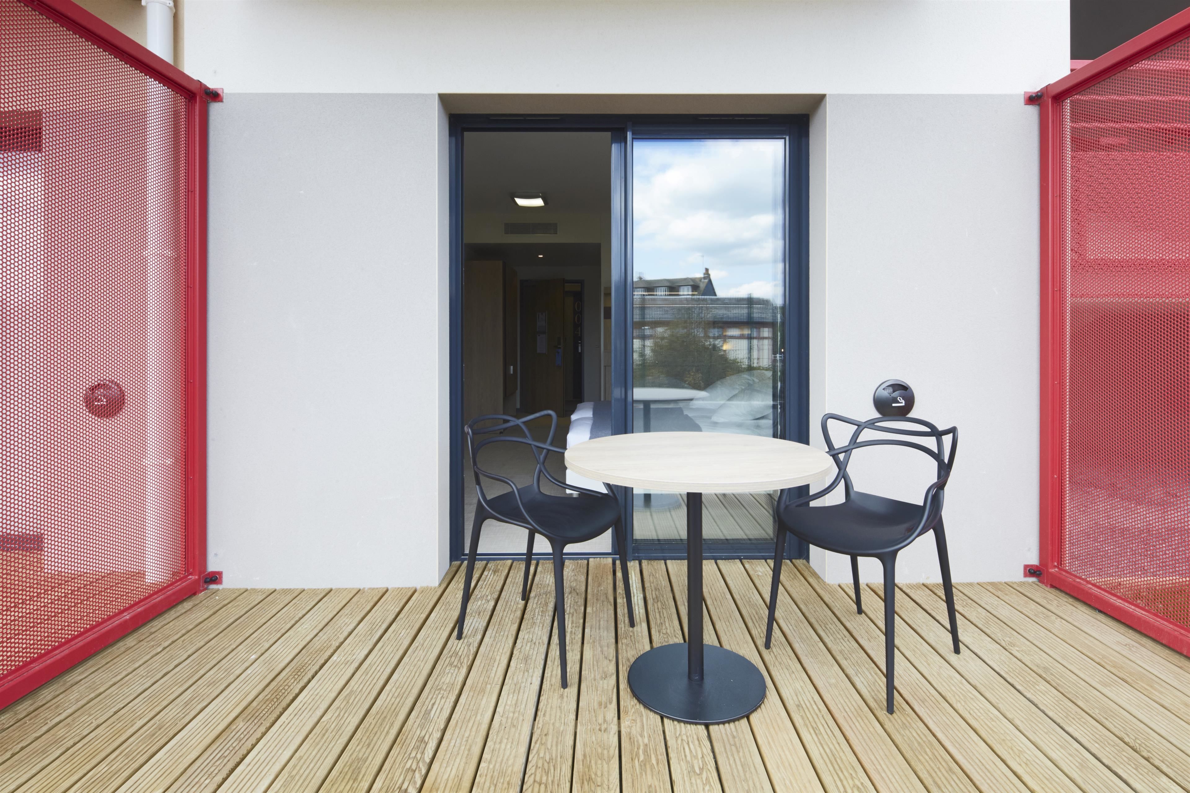Une petite terrasse extérieure avec une table ronde et deux chaises, une porte-fenêtre coulissante et une clôture en treillis rouge.