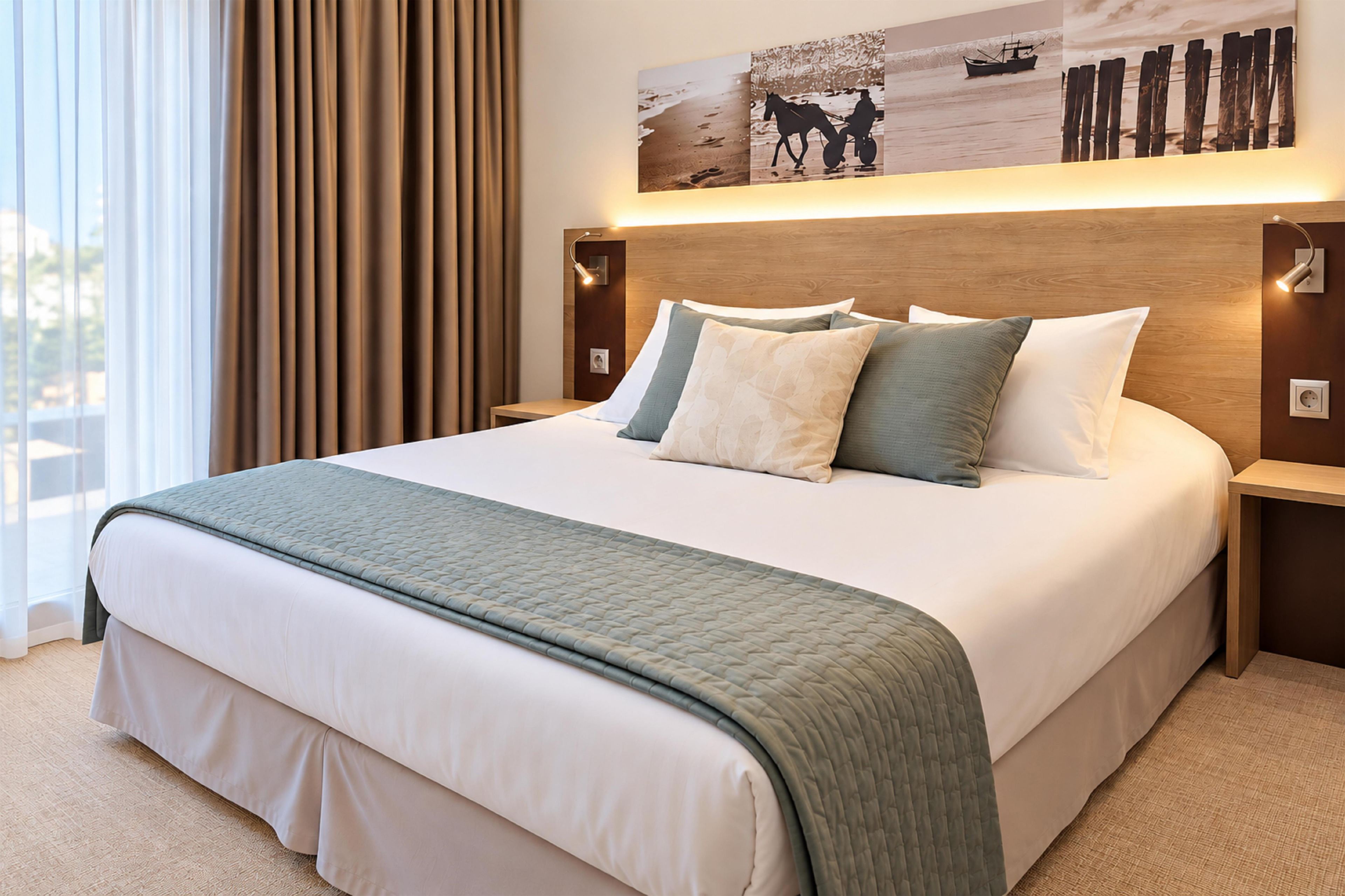 A hotel room with a large bed, a headboard with a light fixture, a nightstand, a wall-mounted artwork, and a view of the outdoors through a window with curtains.