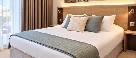 A hotel room with a large bed, a headboard with a light fixture, a nightstand, a wall-mounted artwork, and a view of the outdoors through a window with curtains.