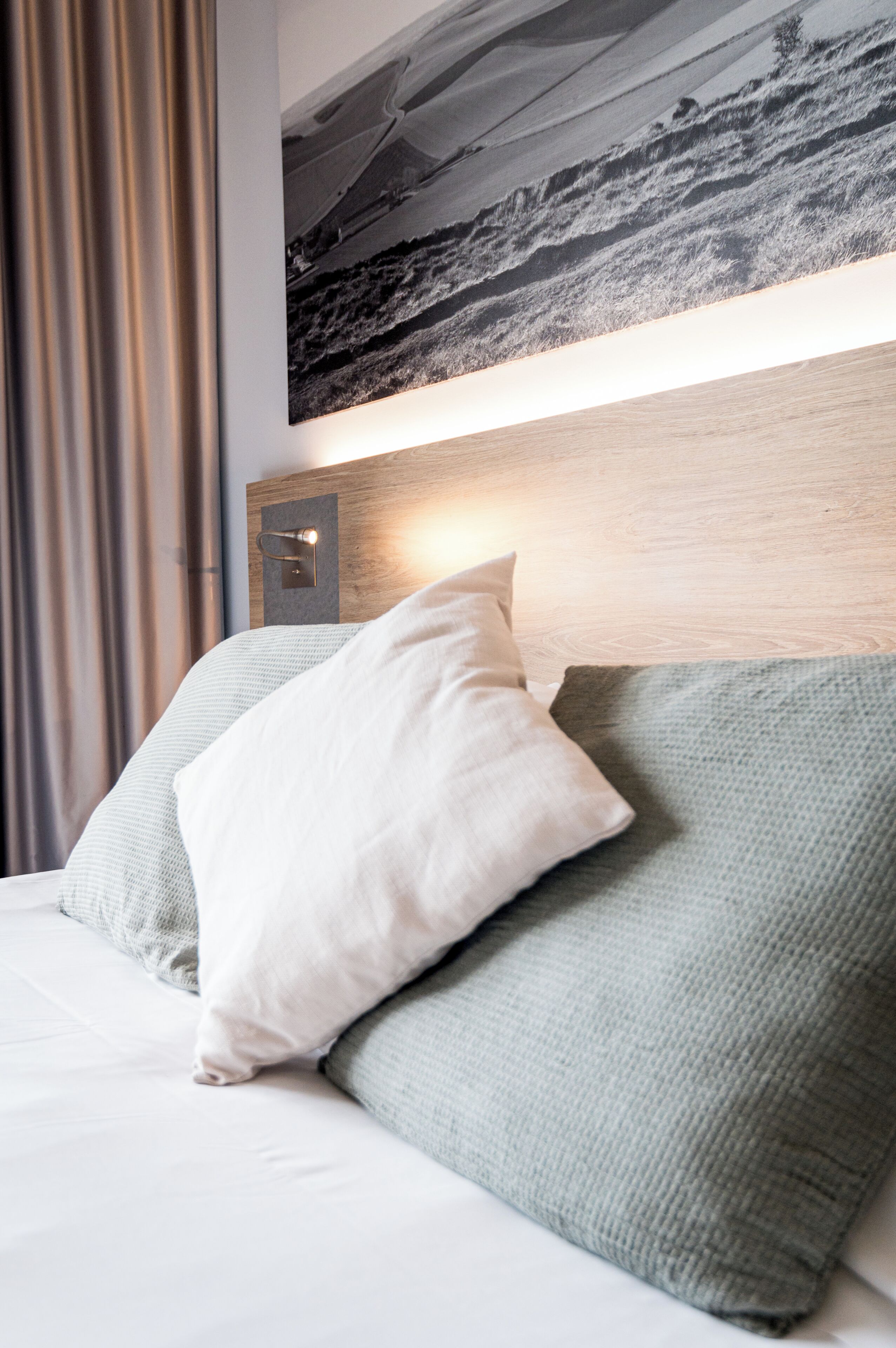 Een opgemaakt bed met twee kussens, een hoofdbord met een lamp en een landschapsschilderij erboven.