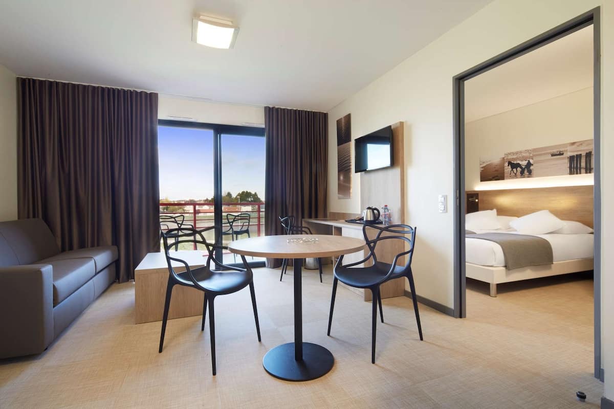 Una habitación de hotel moderna con sofá, una mesa redonda, sillas, una cama, un televisor y un balcón con cortinas.