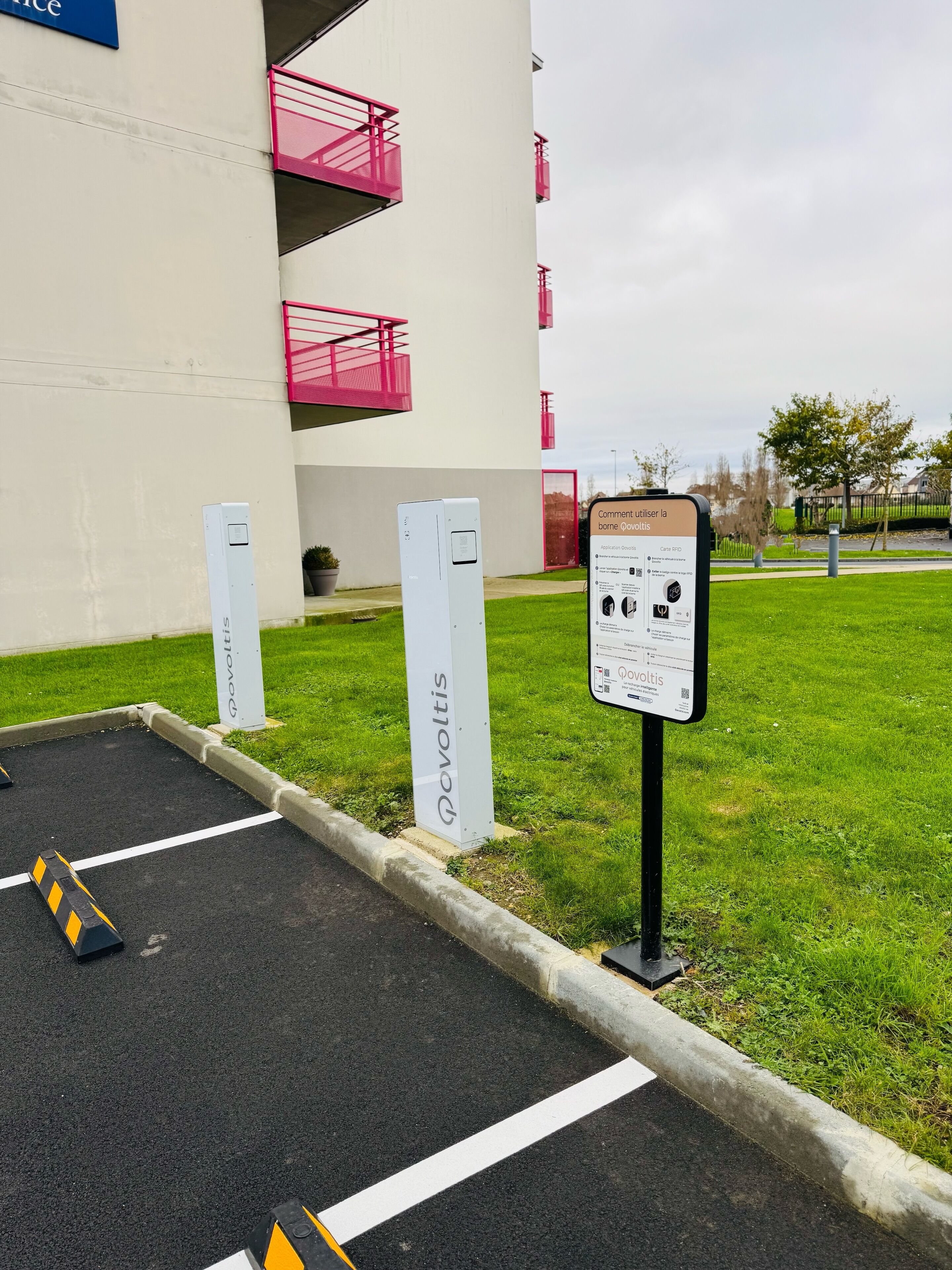Un espace de stationnement équipé de bornes de recharge pour véhicules électriques, un panneau d’information et un bâtiment doté de balcons.