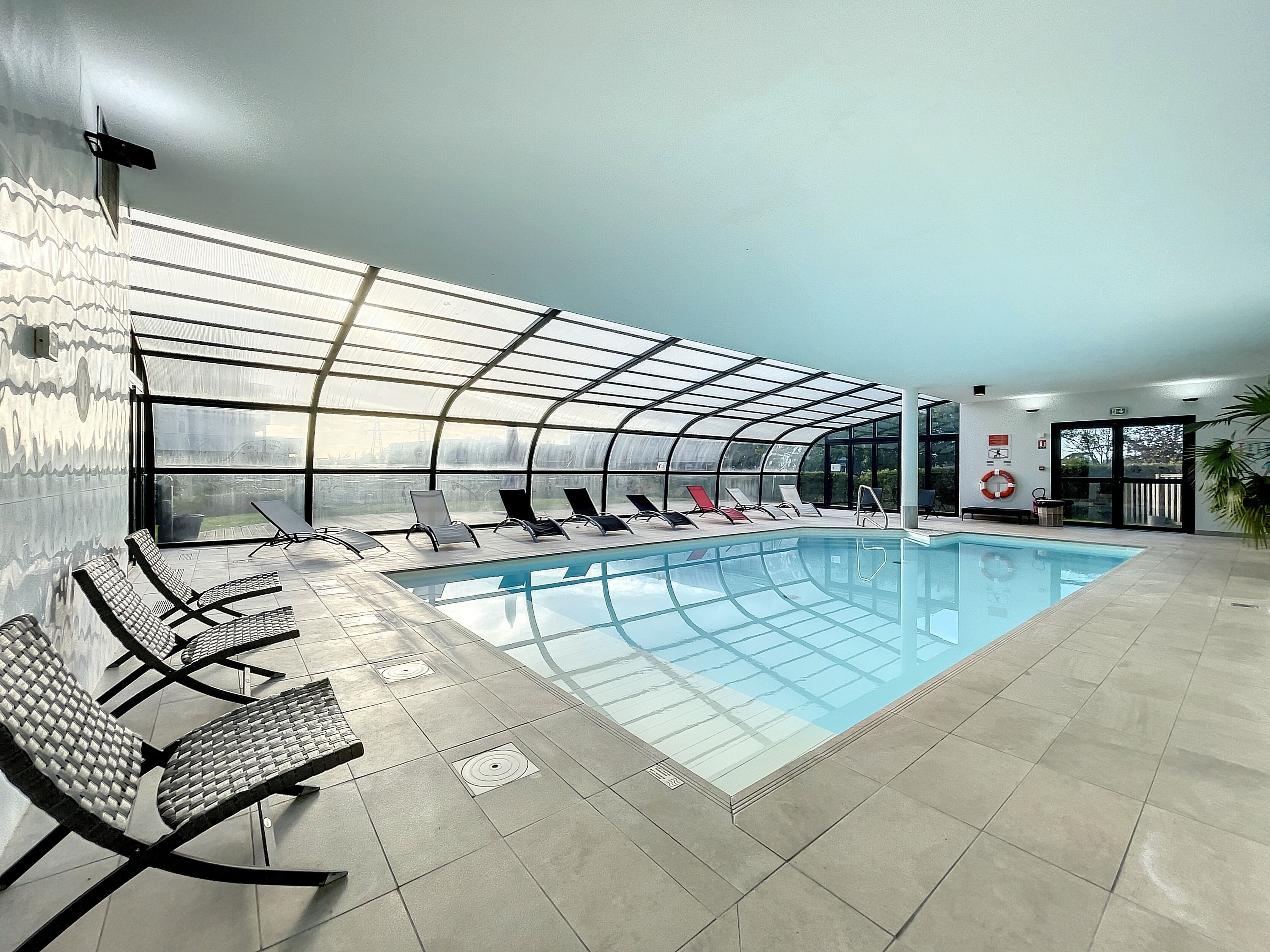 Une piscine intérieure dotée d’un toit en verre, de chaises longues et offrant une vue sur l’extérieur.