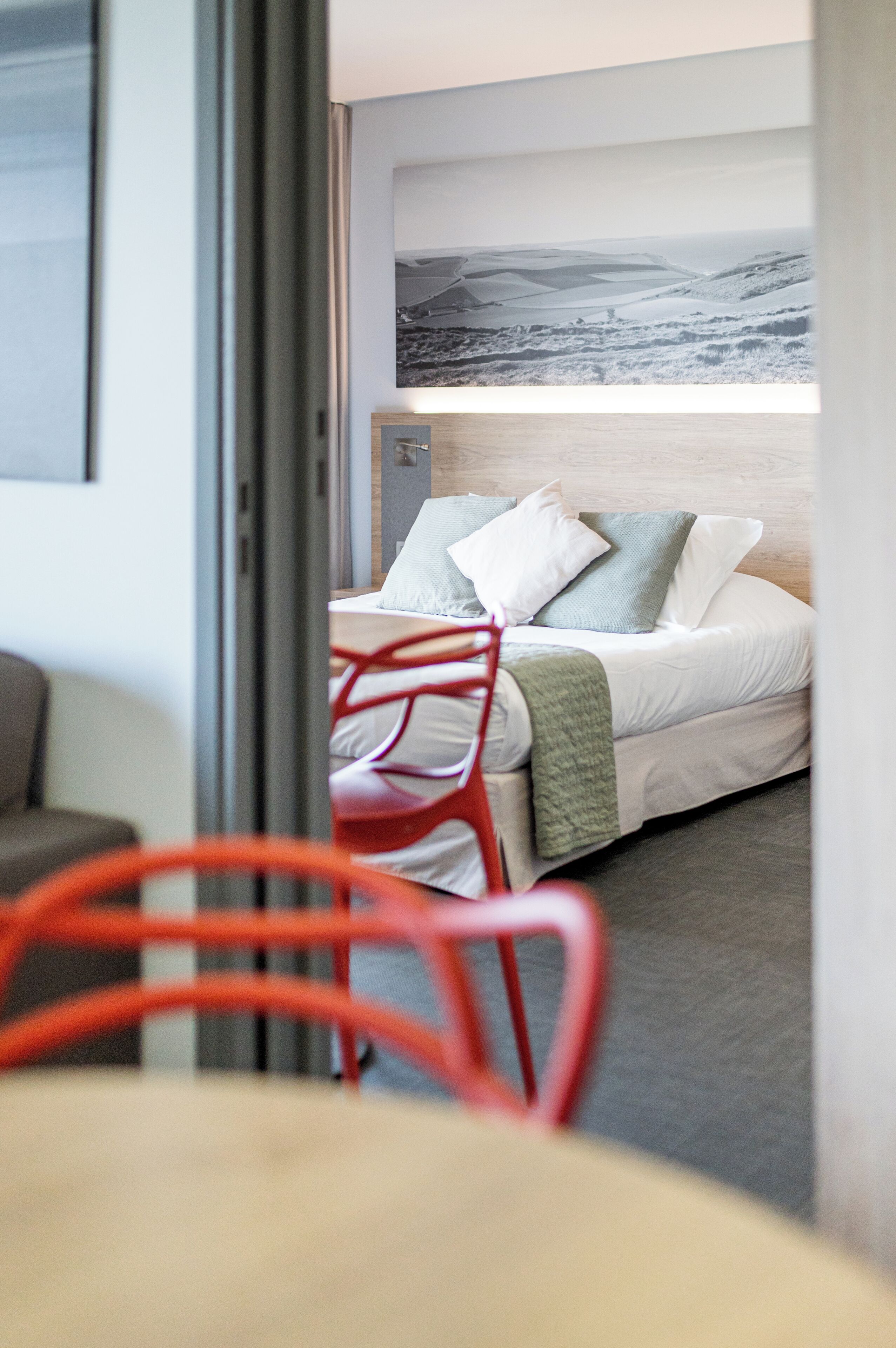 A hotel room with a bed, a red chair, a table, and a painting on the wall.