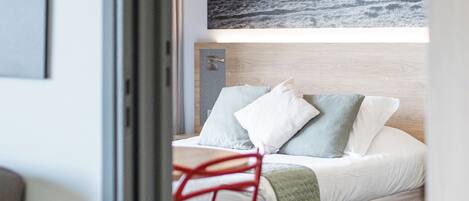A hotel room with a bed, a red chair, a table, and a painting on the wall.