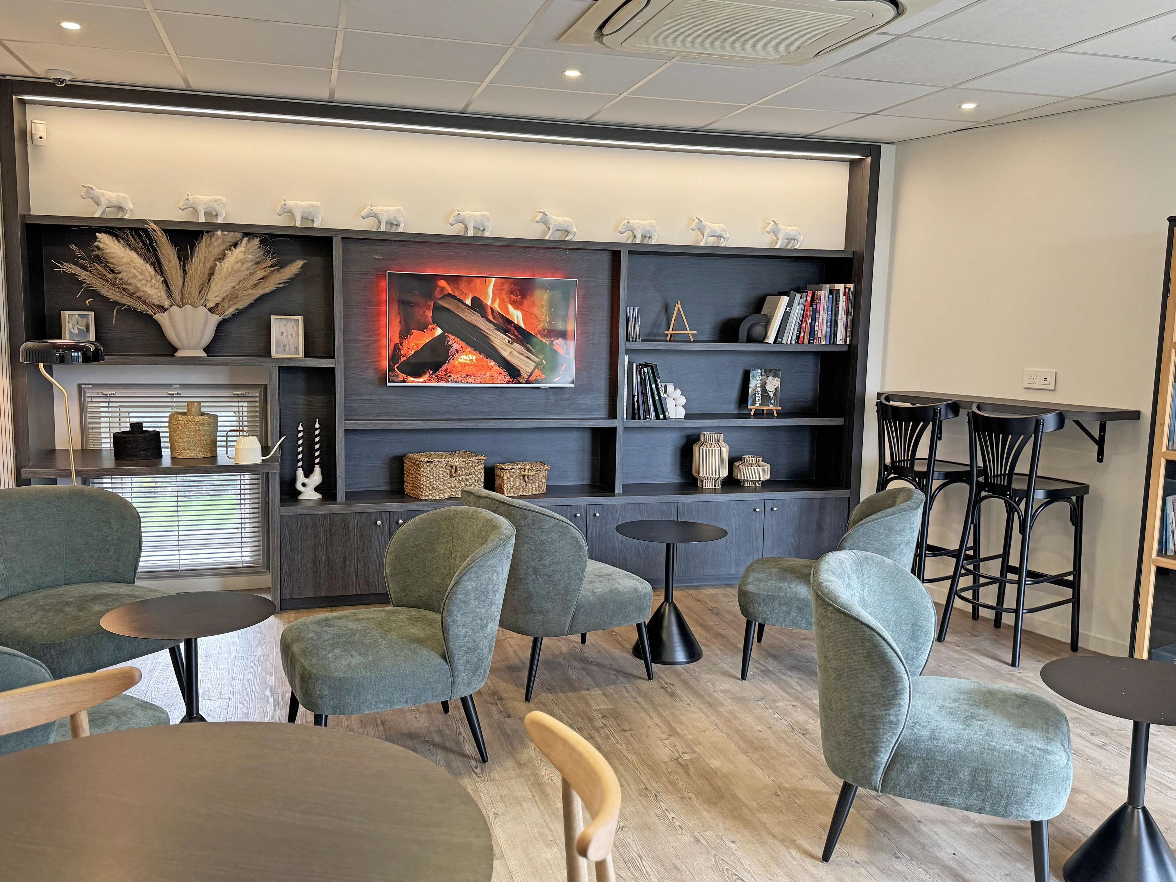 A modern lounge area with grey upholstered chairs, a fireplace with a framed artwork, and a bookshelf with decorative items.