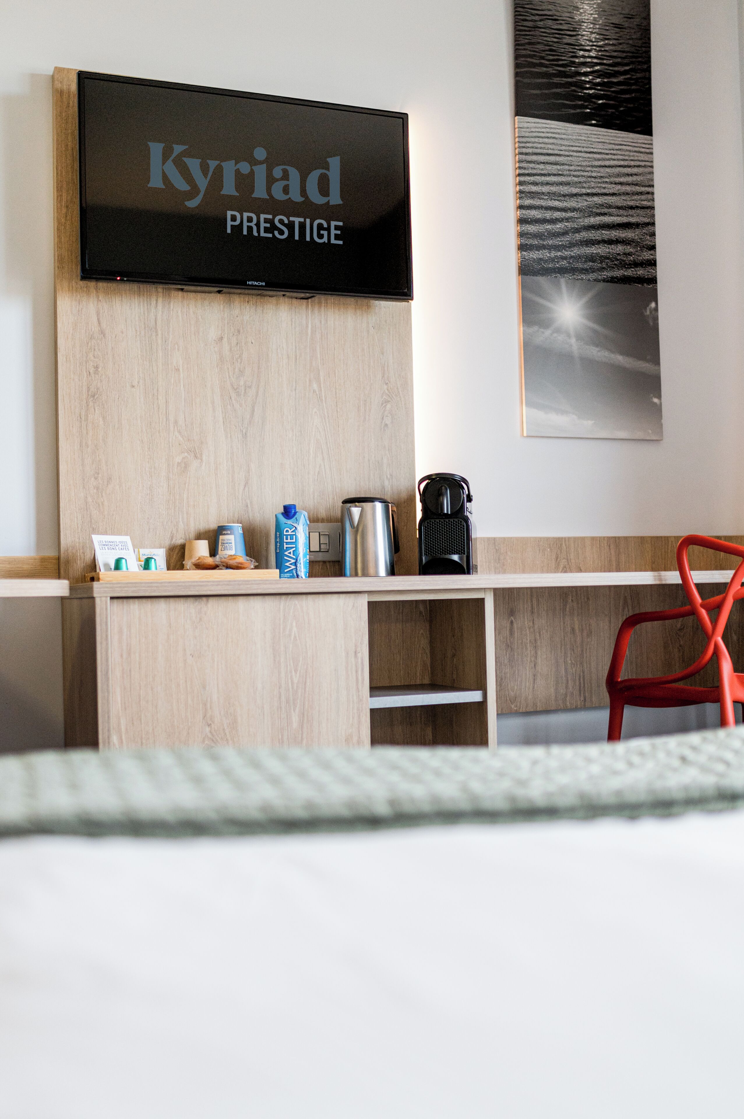 Habitación de hotel con televisor de pantalla plana mostrando 'Kyriad Prestige', un escritorio de madera con cafetera, botella de agua y una fotografía enmarco de una playa.