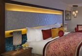 A hotel room with a large bed, a wooden nightstand, a decorative headboard, and a patterned throw blanket.