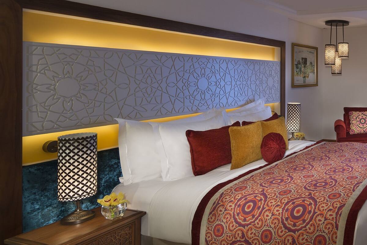 Une chambre d’hôtel avec un grand lit, une table de chevet en bois, une tête de lit décorative et un plaid à motifs.