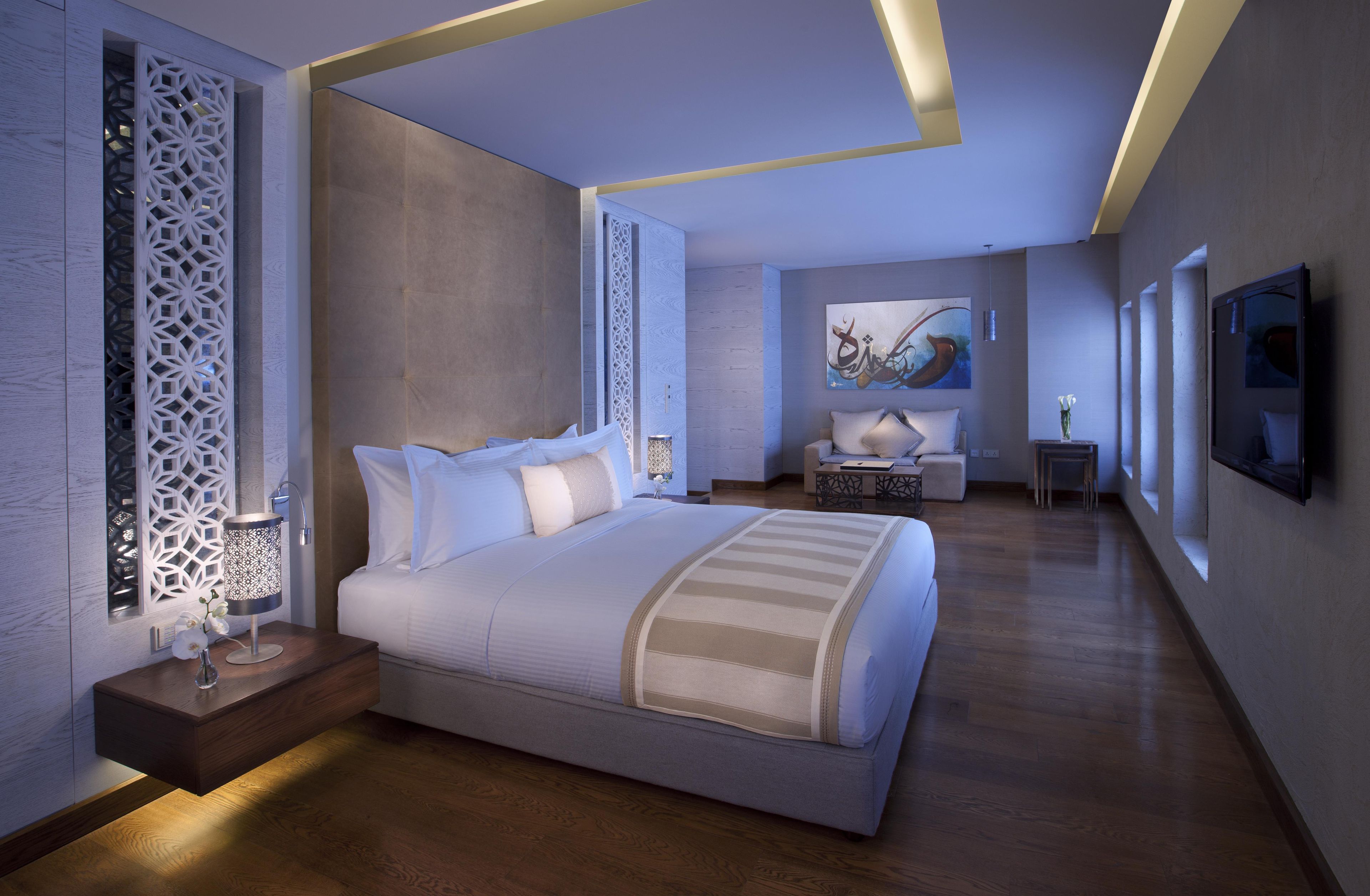 A modern bedroom with a large bed, a decorative wall panel, a small table with a lamp, and a painting on the wall.
