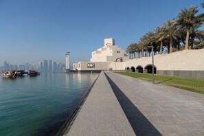 A waterfront walkway with a modern building, palm trees, and boats. - Souq Waqif Boutique Hotels by Tivoli (Doha)