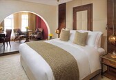 A hotel room with a large bed, a wooden nightstand, a mirror, and a view through an arched doorway to another room with a desk and chairs.