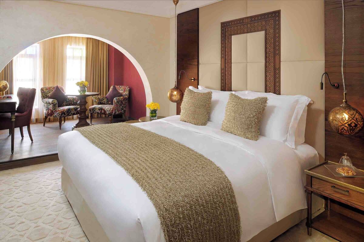 Une chambre d’hôtel avec un grand lit, une table de chevet en bois, un miroir et une vue sur une autre pièce, accessible par un passage en arc, où se trouvent un bureau et des chaises.