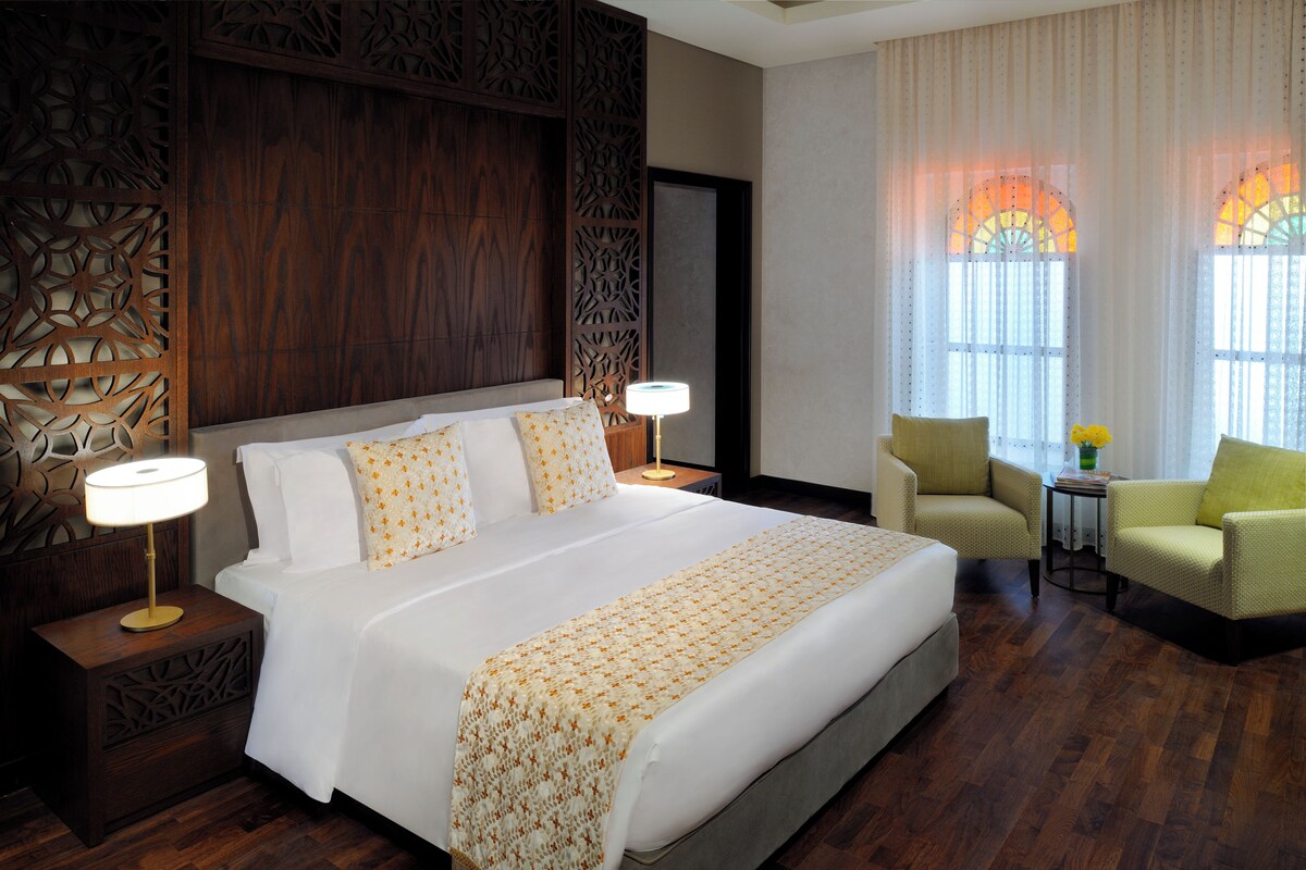 A hotel room with a large bed, two bedside lamps, a chair, a small table, and a decorative wall panel.