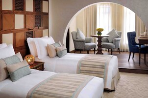 Habitación de hotel con dos camas, un separador de madera, una mesita con un florero y un sillón azul.