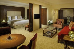 A hotel room with a large bed, a sitting area with a red sofa and armchairs, a coffee table, and a dining table with a vase of flowers.