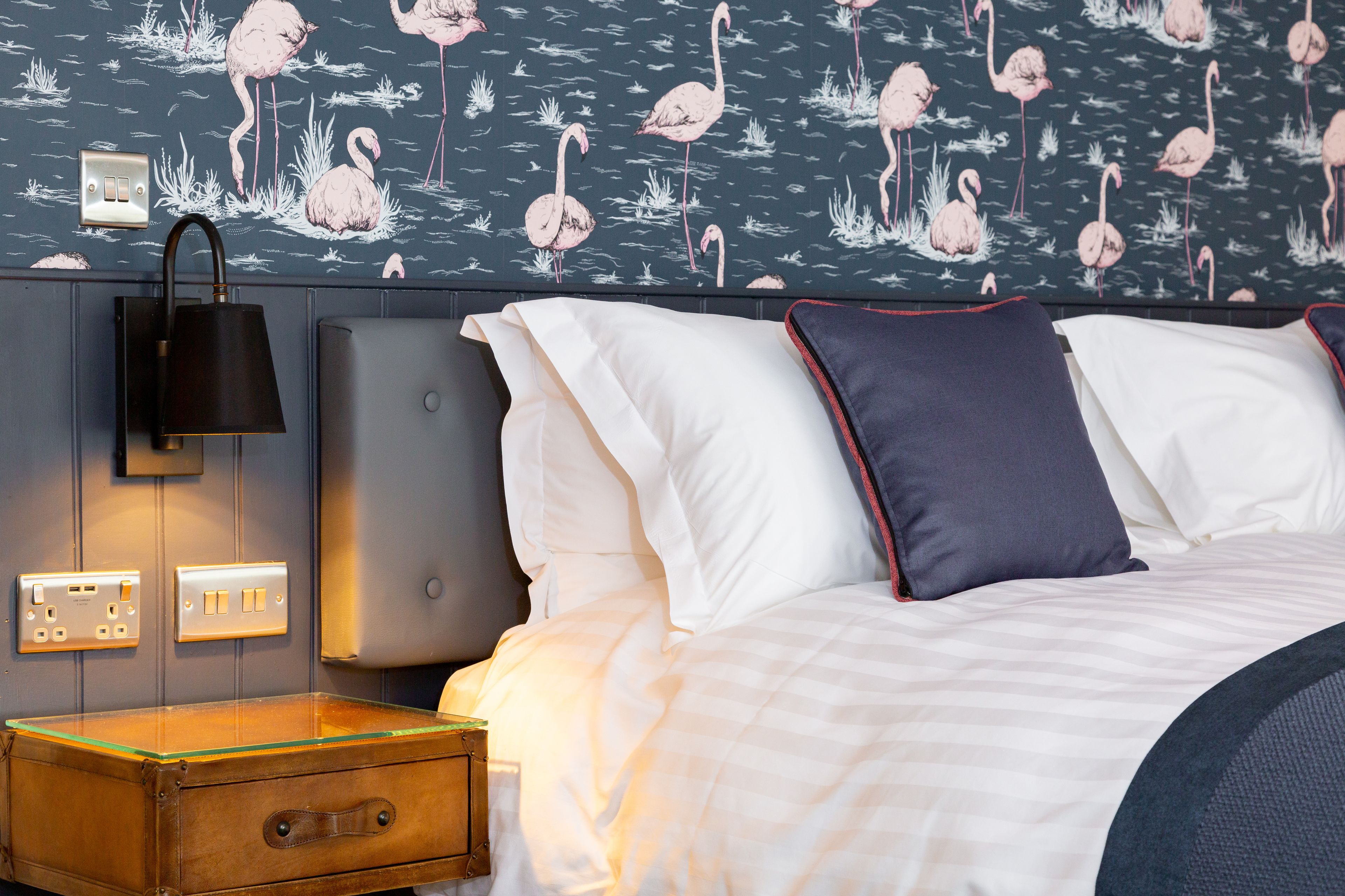 A hotel room with a bed, a nightstand, a lamp, and a wall with a flamingo pattern.