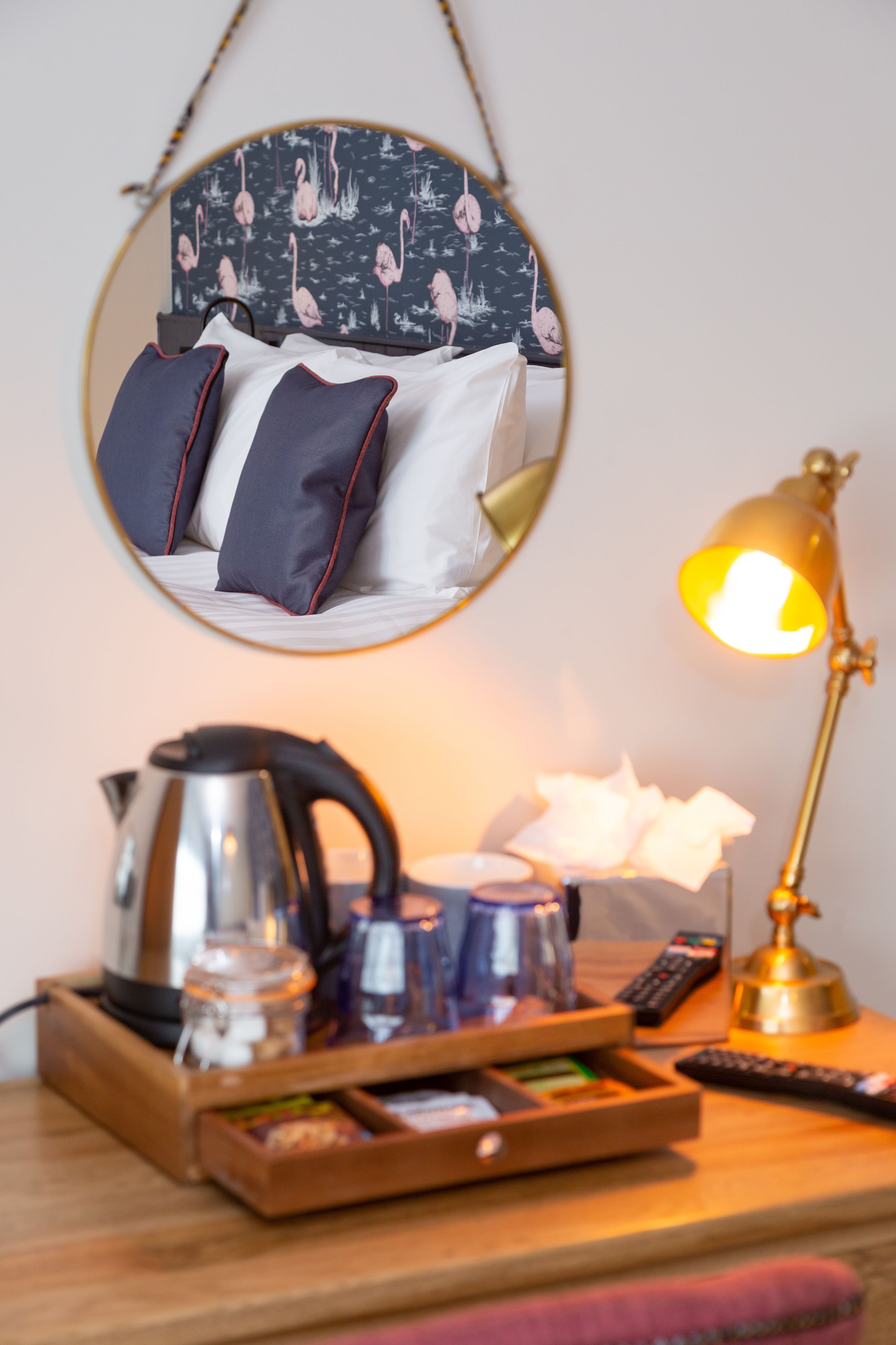 Habitación de hotel impecable con una cama, una lámpara de noche, un reposabajares con una tetera, vasos y un control remoto.