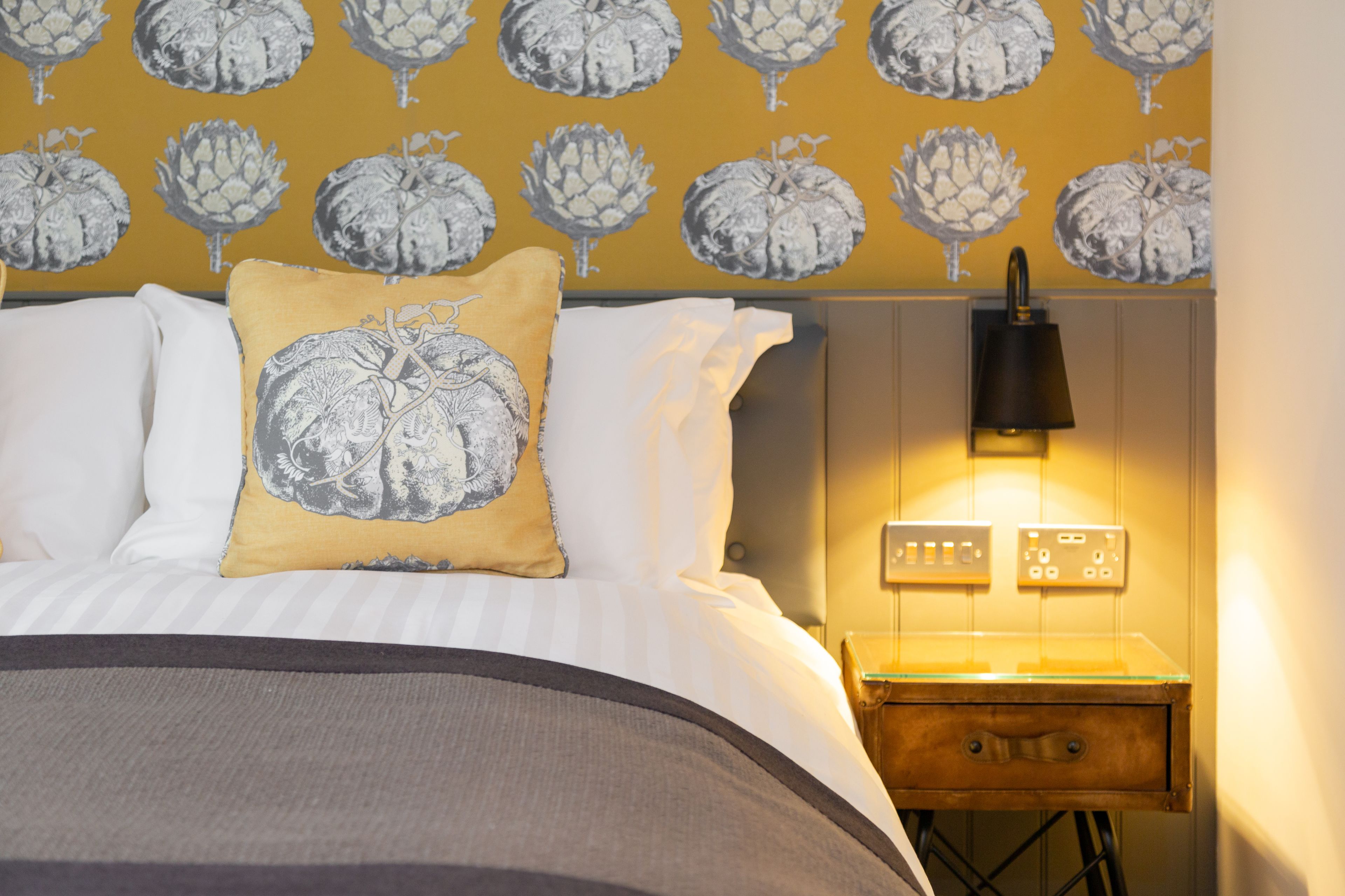 A neatly made bed with striped bedding, a decorative pillow, a bedside table with a lamp, and a wall with a repeating art print.