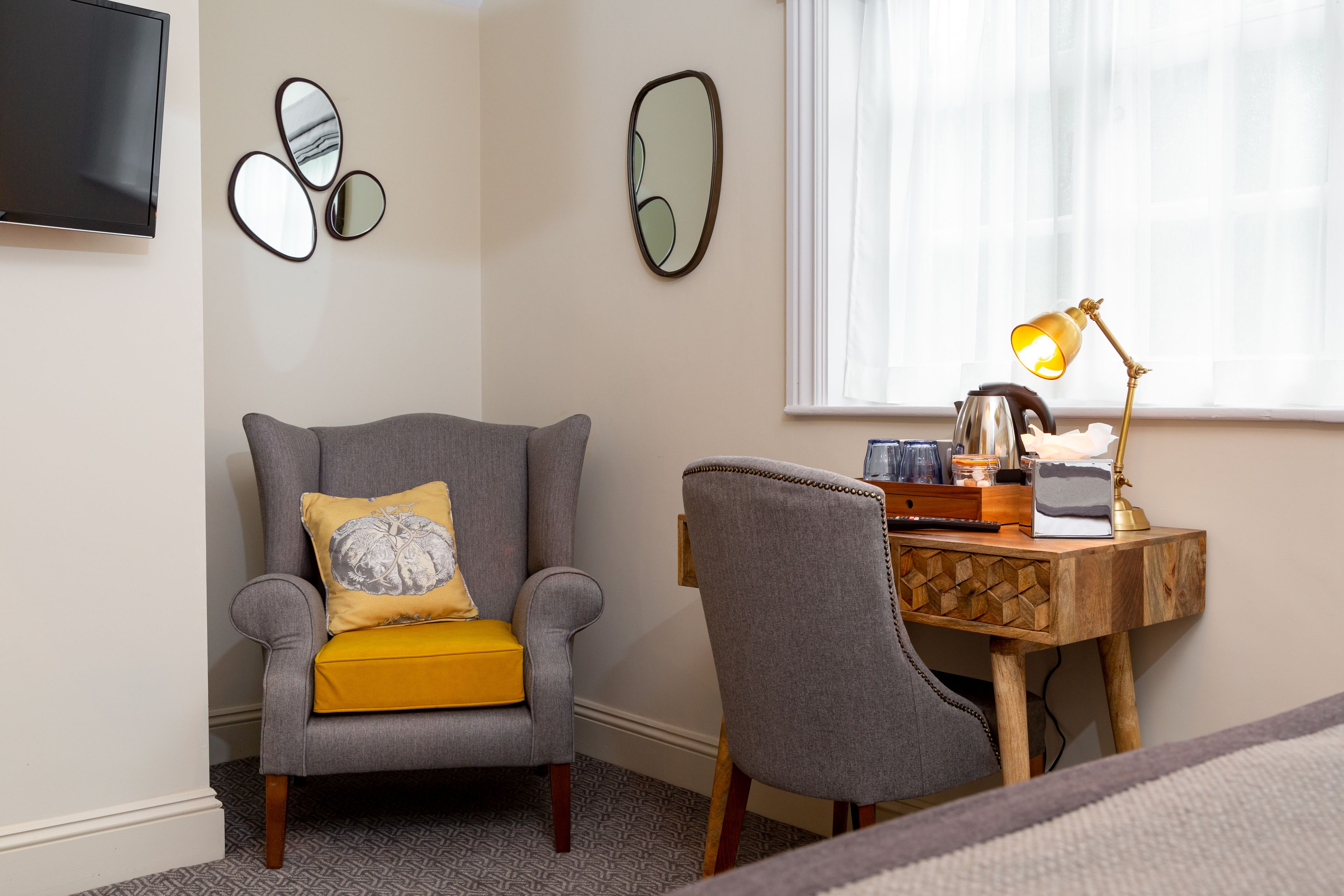 A room with a grey armchair with a yellow cushion, a wooden side table with a lamp, and a TV mounted on the wall.