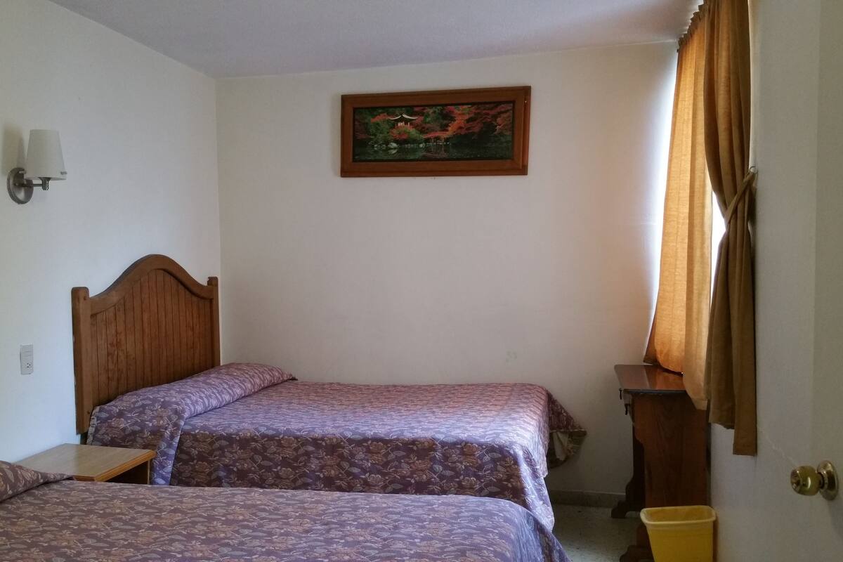 A room with two wooden beds, a wall-mounted light, a painting, and a door with curtains.