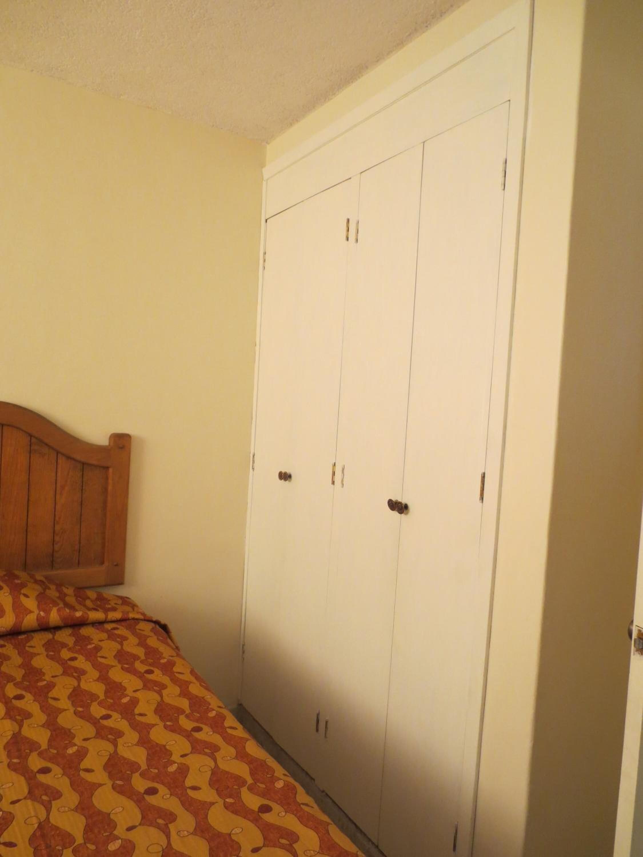 Un dormitorio con cama, cabecera de madera y un amplio armario blanco de dos puertas y dos cajones.