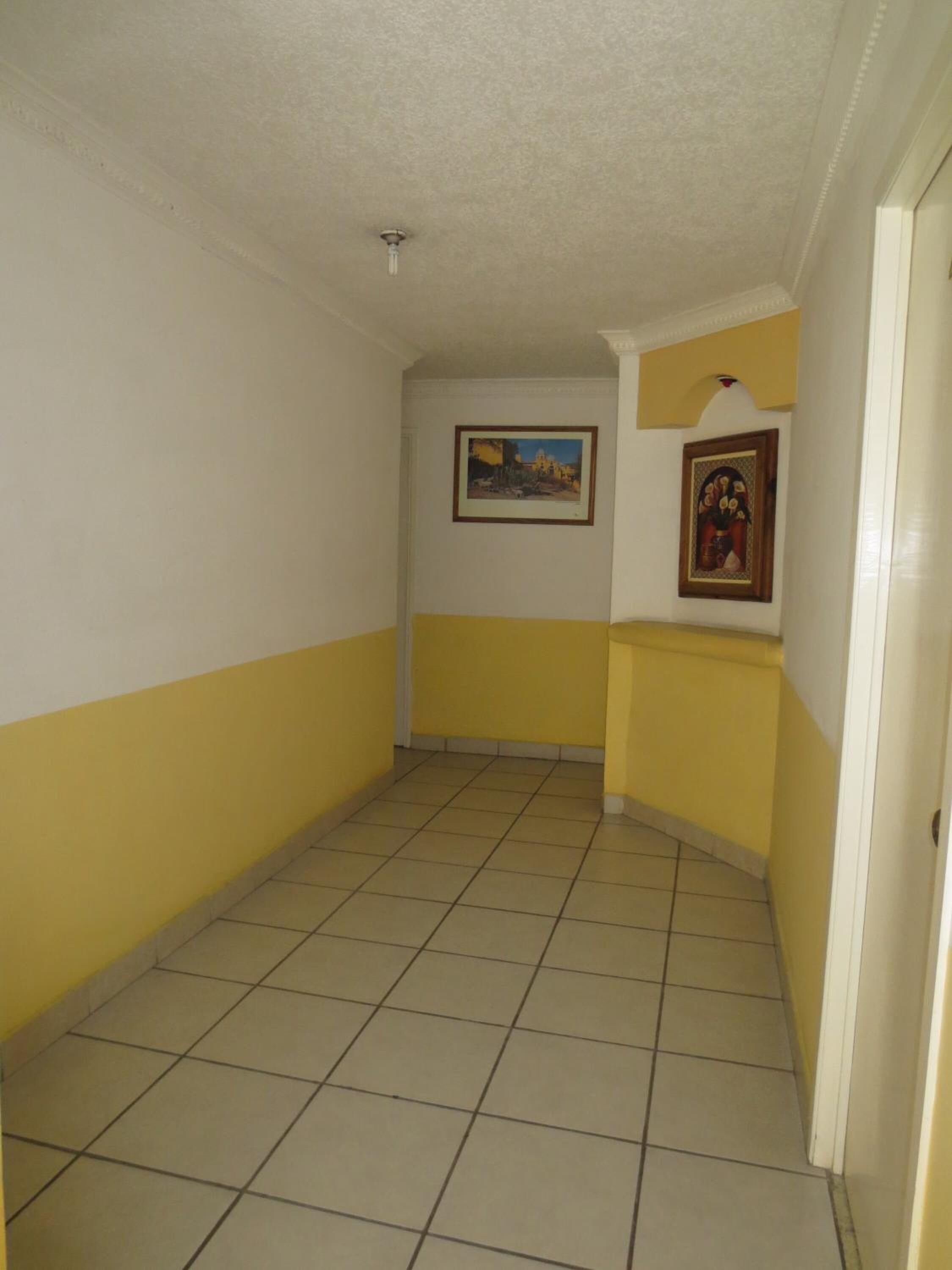 Un pasillo estrecho con paredes de color amarillo y blanco, suelo de baldosas y dos cuadros enmarcados.