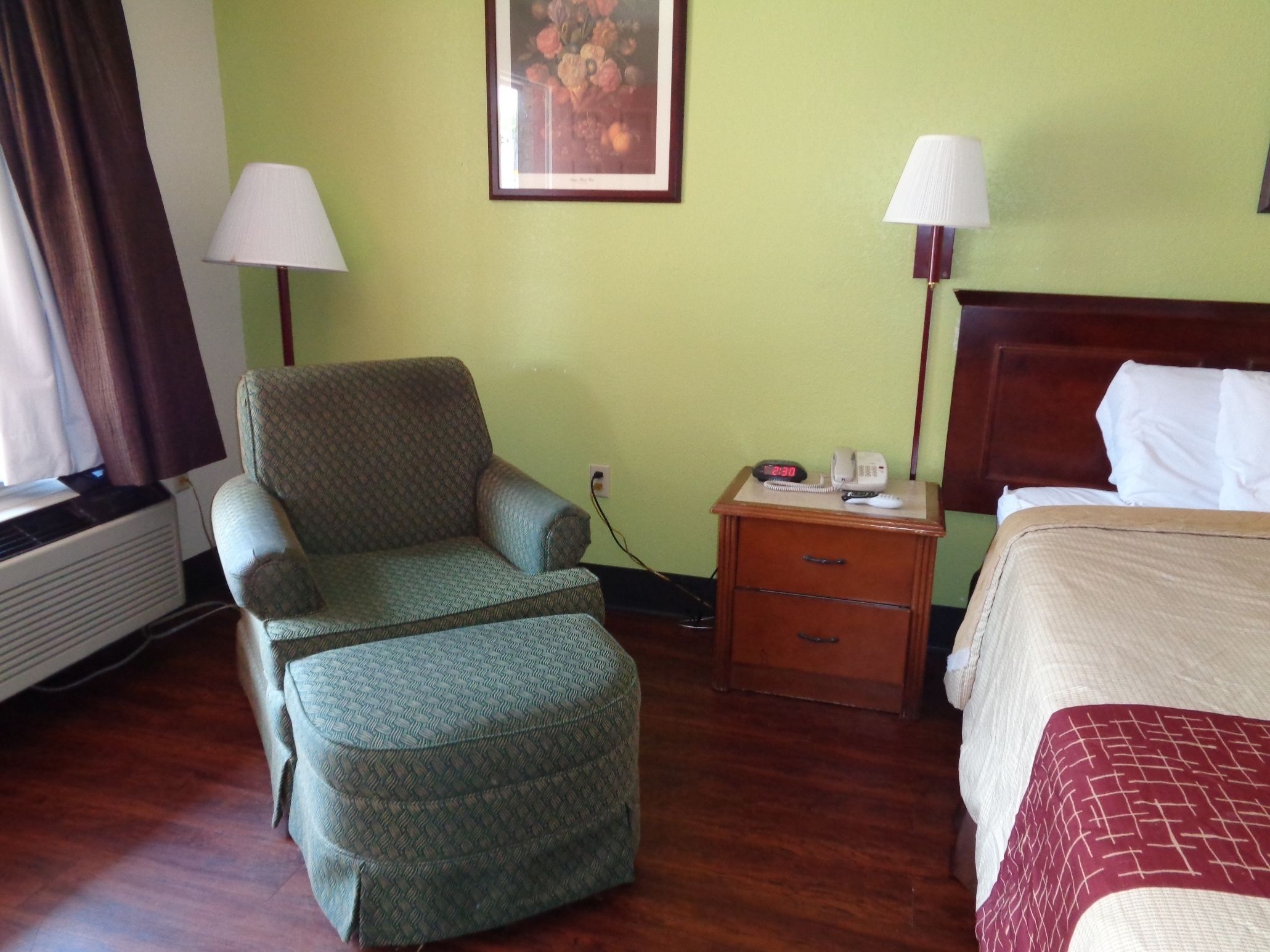 Ein Hotelzimmer mit grüner Wand, einem hölzernen Nachttisch mit Lampe, einem Sessel, einem Bett mit rot-weißer Bettdecke und einem gerahmten Bild an der Wand.