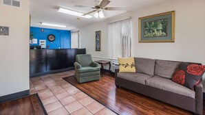 Una zona de recepción con pared azul, un mostrador de recepción, un sillón verde, un sofá marrón con cojines rojos y amarillos, y un ventilador de techo.